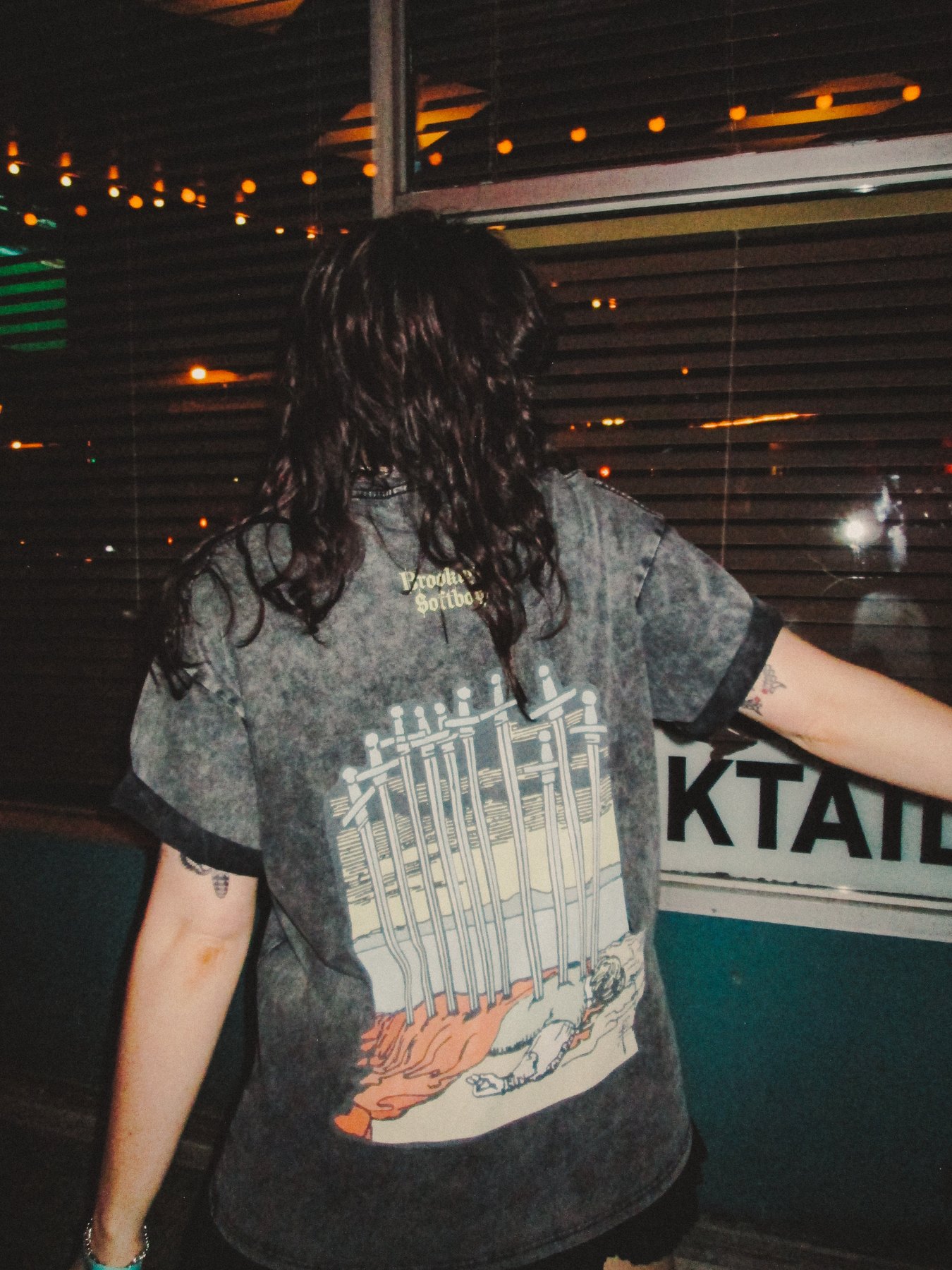 Model from behind in front of a neon-lit storefront, showing the Ten of Swords back print.