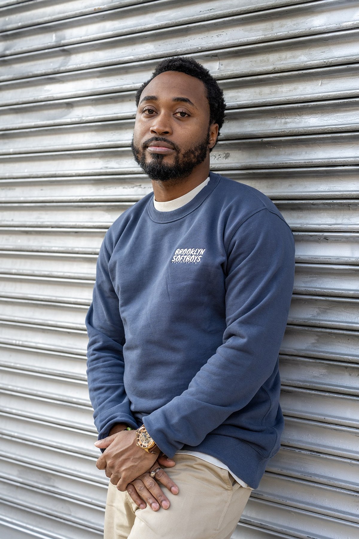 Man in a navy SoftBoys crewneck leaning against a steel roll-down gate, looking at the camera.
