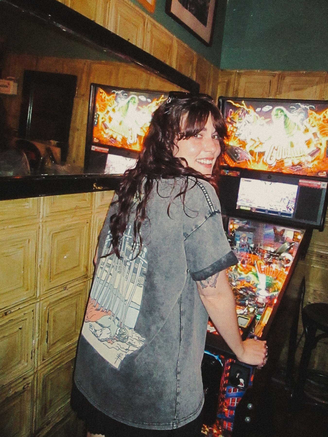 Model at a pinball machine in a dim bar, wearing the mineral-washed Ten of Swords tee.