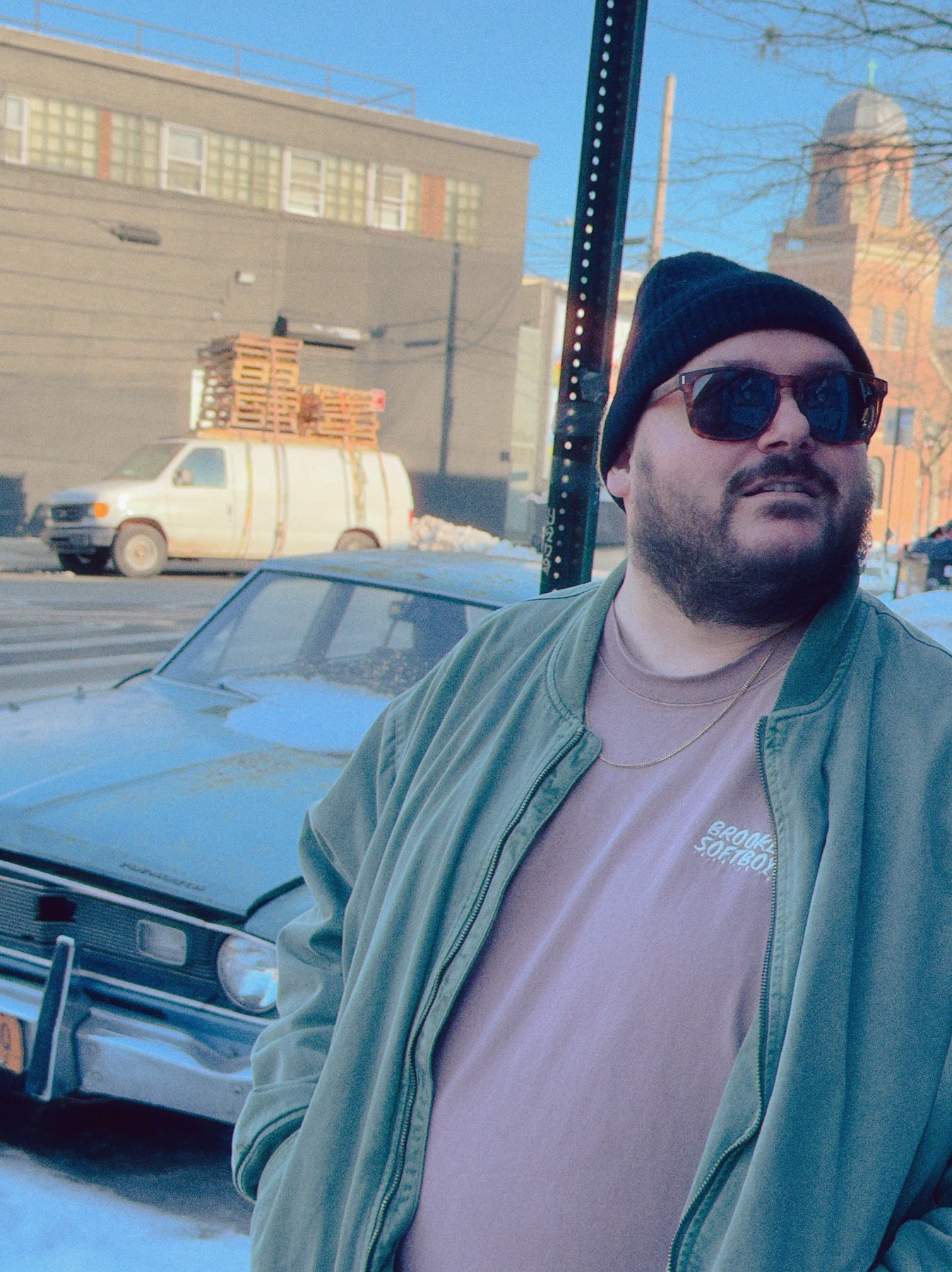 Man in sunglasses, beanie and a green bomber jacket beside a vintage car in the snow, wearing a mauve SoftBoys tee.