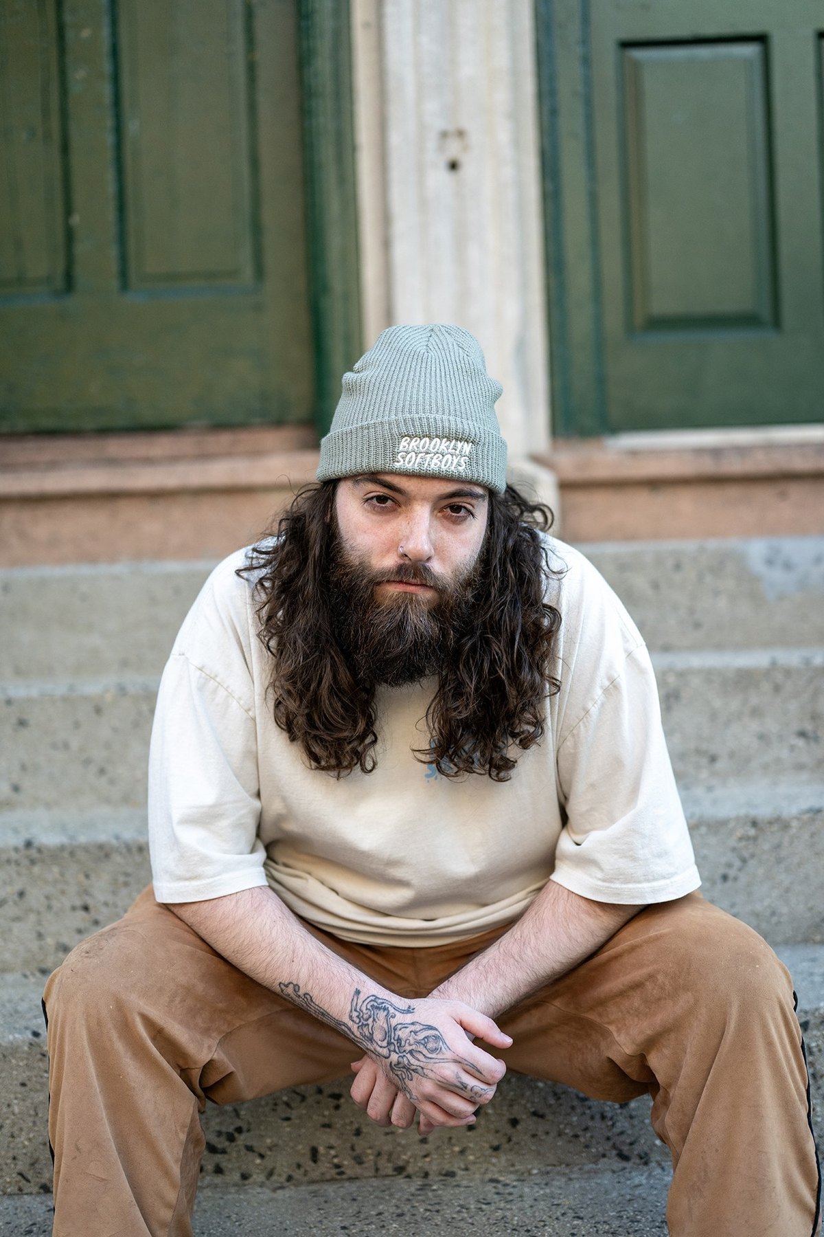 Man in a cream tee and sage beanie sitting on stoop steps, hands clasped.