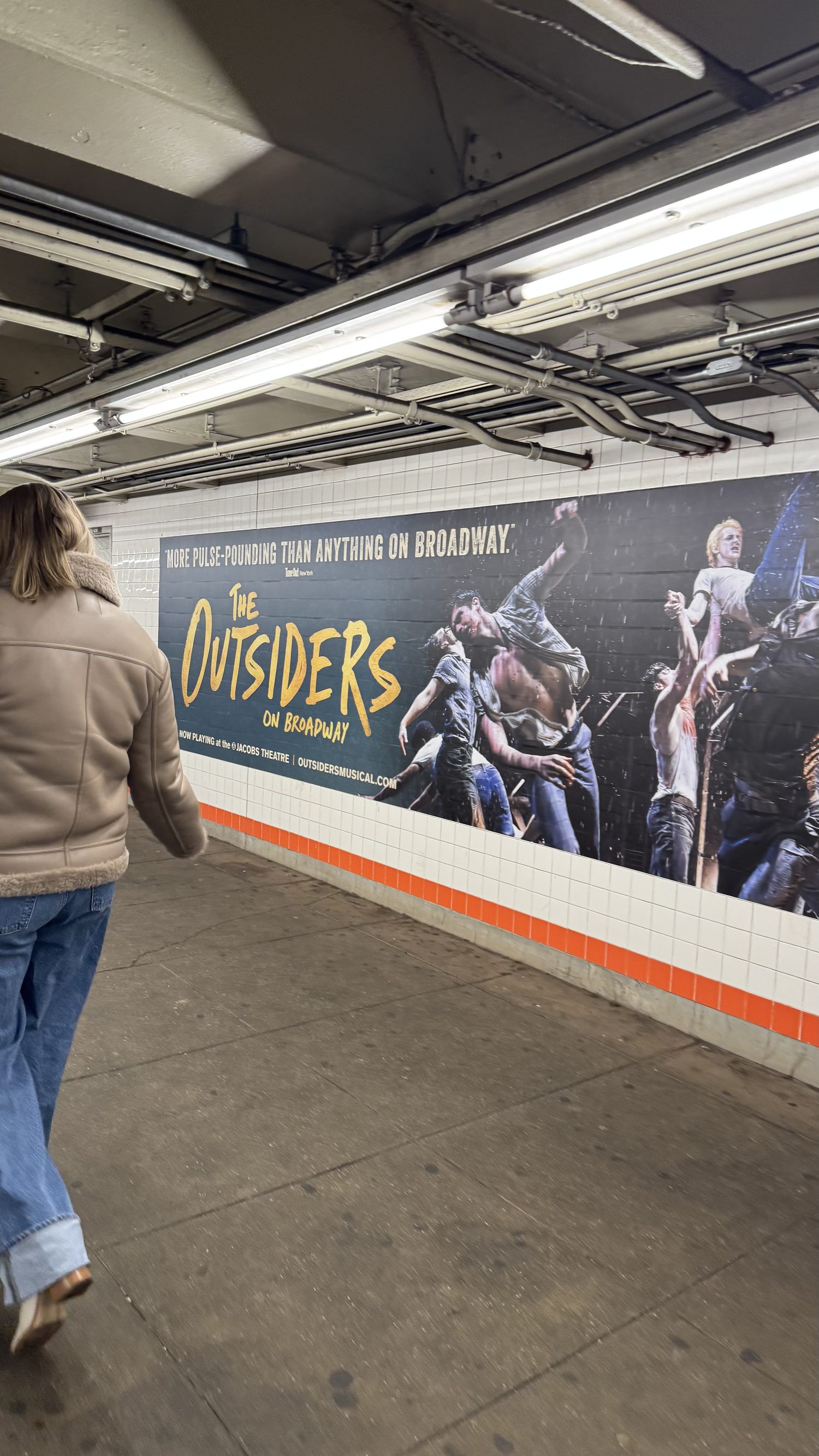 A commuter walking past a long tiled wall banner with the Outsiders title and the cast.