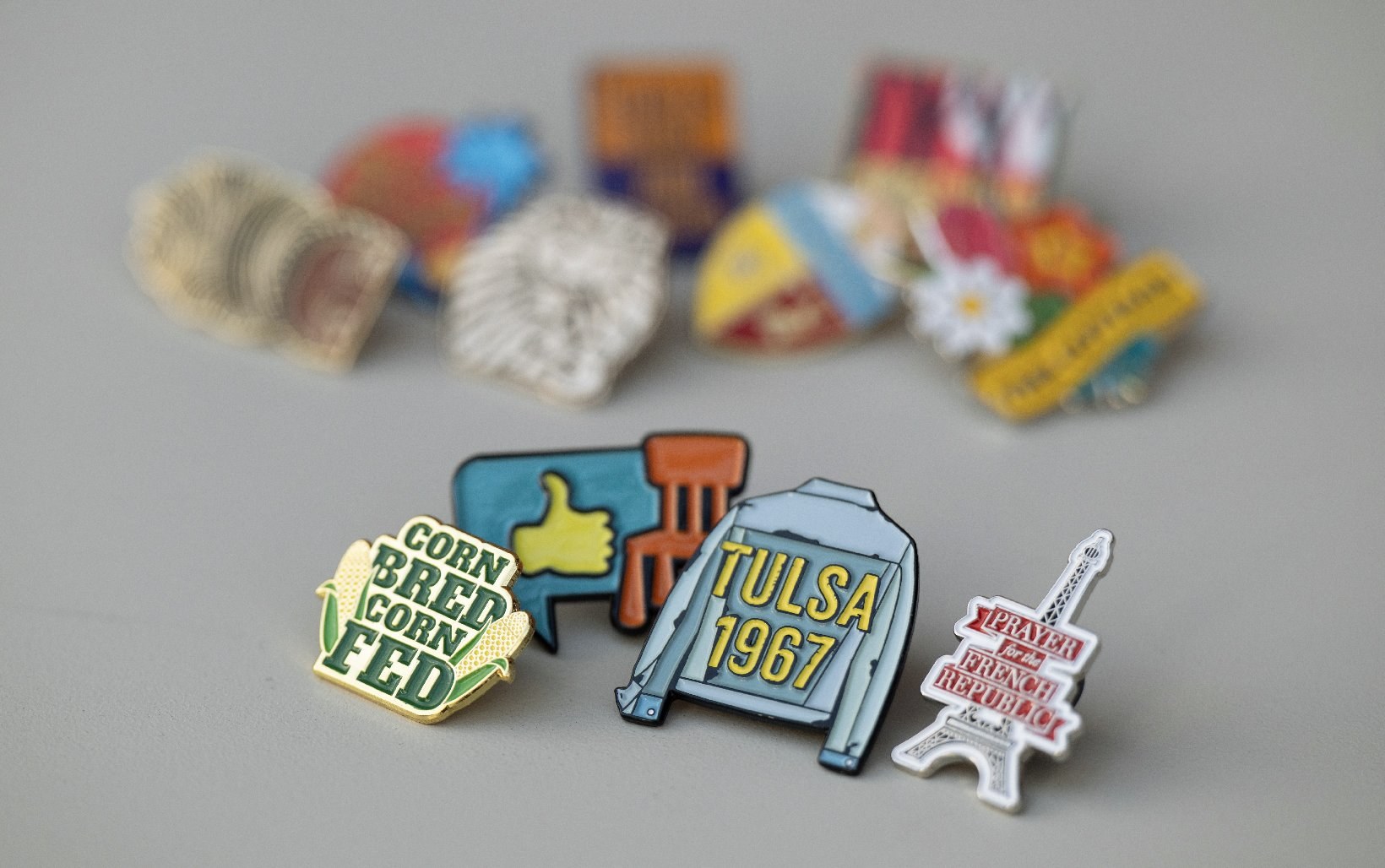 A group of enamel pins for Broadway shows on a gray surface — Corn Bred Corn Fed, a thumbs-up speech bubble with a chair, a Tulsa 1967 denim jacket, and an Eiffel Tower — with more pins blurred behind.