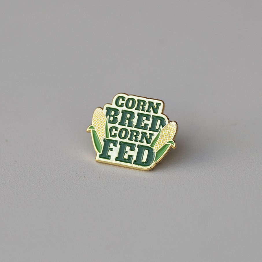 Enamel pin with the words Corn Bred Corn Fed between two ears of corn, for Shucked.