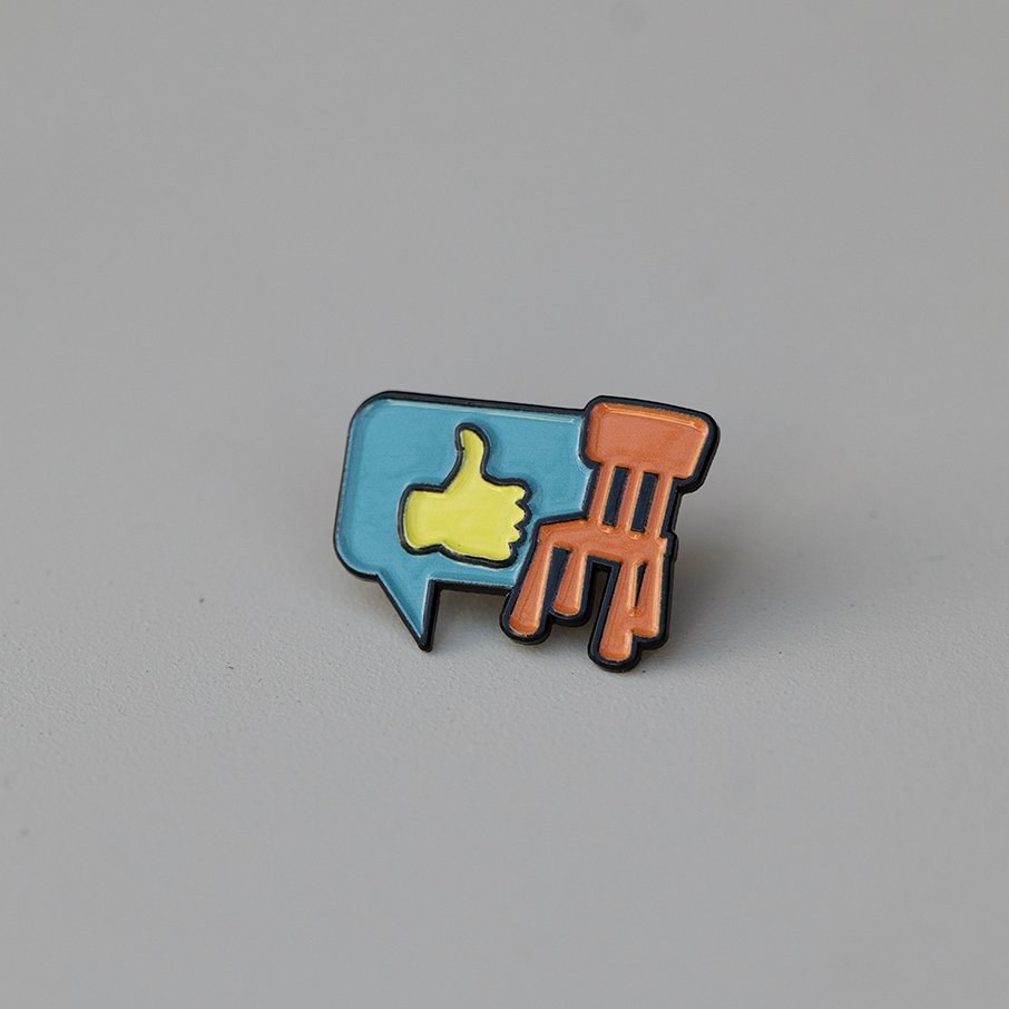 Enamel pin of a blue speech bubble with a yellow thumbs-up and an orange chair, for Eureka Day.