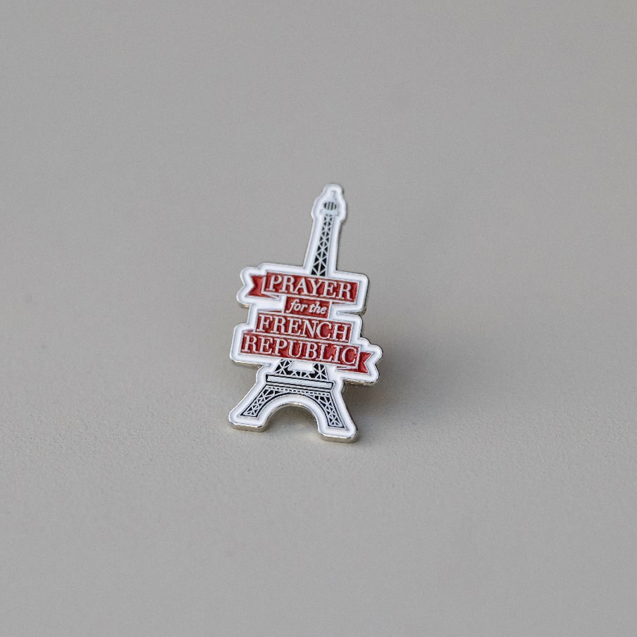 Enamel pin shaped like the Eiffel Tower wrapped in a red banner reading Prayer for the French Republic.
