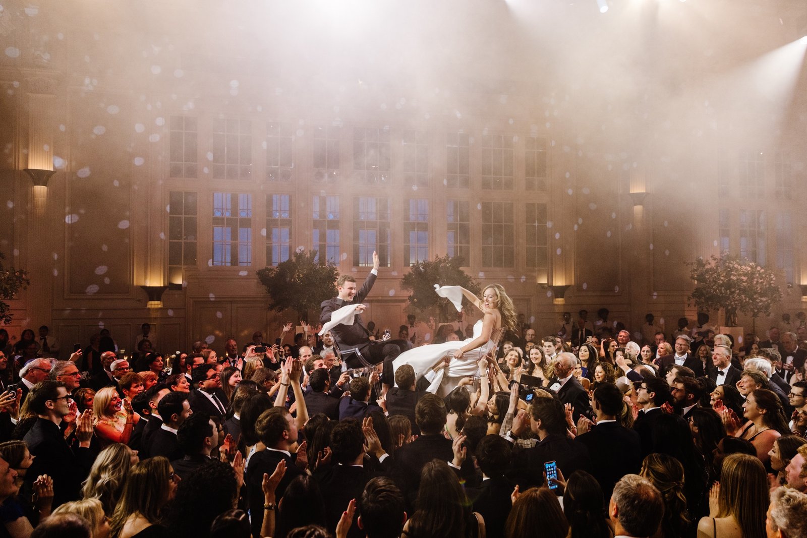 Celebratory hora chair-lift at a wedding reception, warm light and confetti.